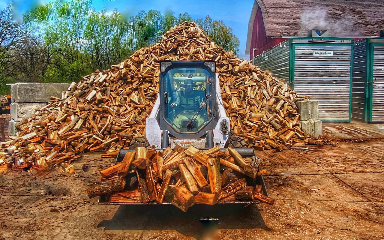 Picture of a firewood being scooped by a bobcat in front of a wood pile.