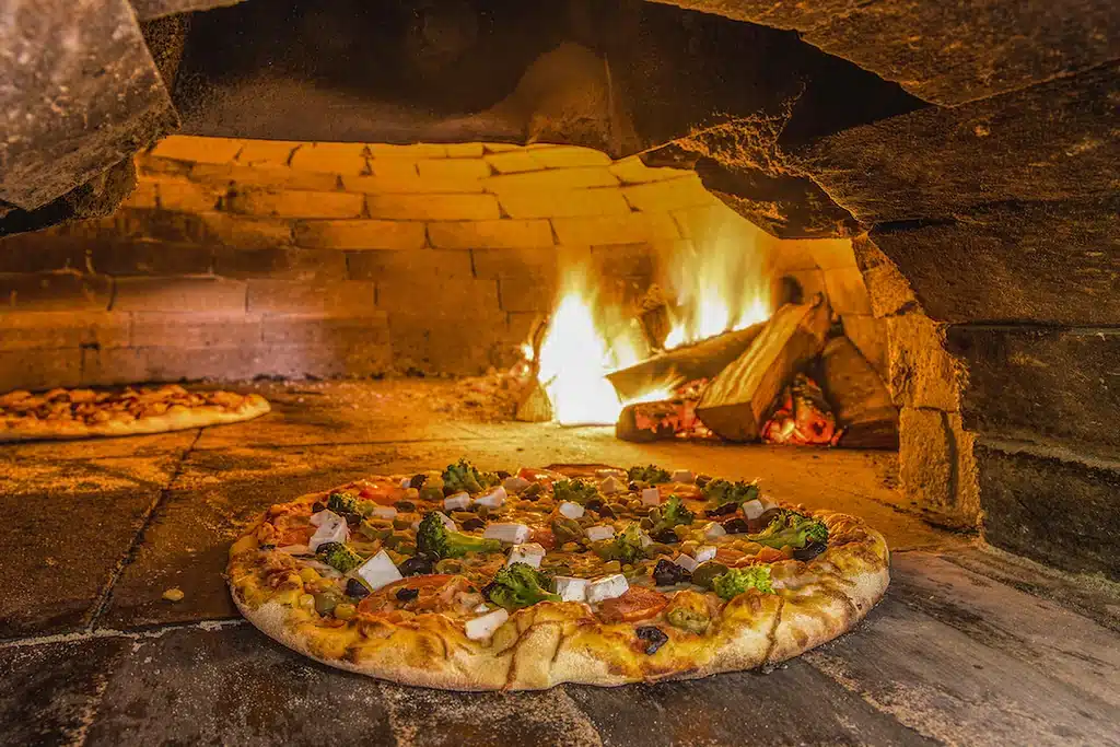Pizza in a brick wood-fired oven, with burning logs and flames at rear right and another pizza at left.