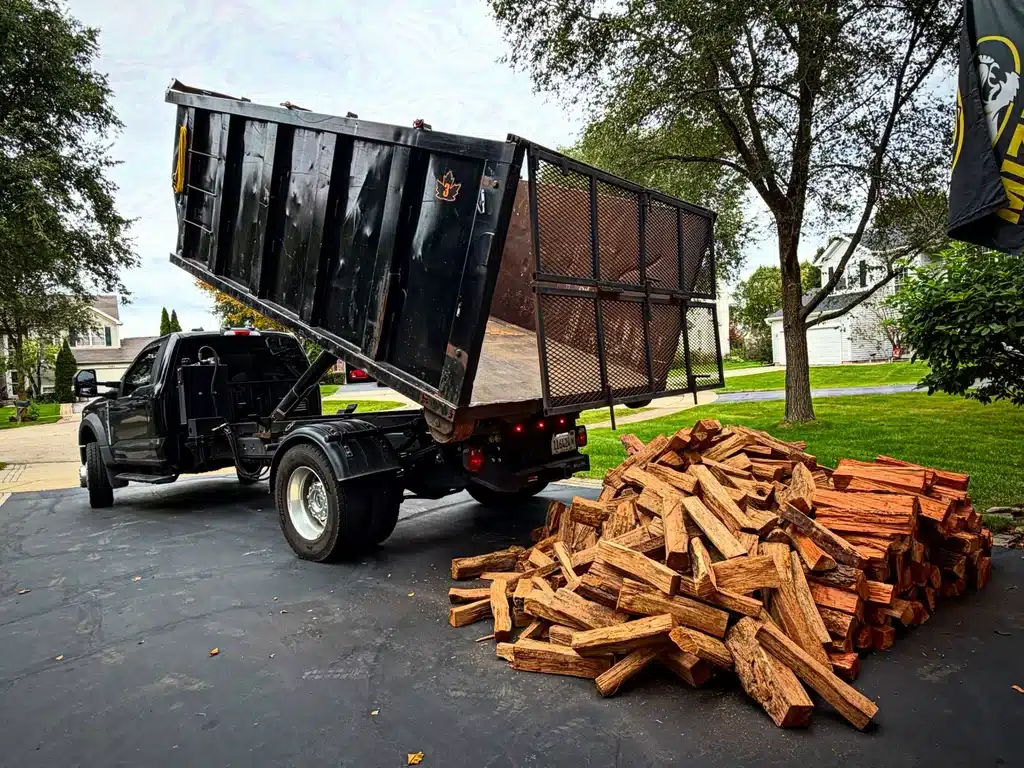 Black dump truck with its bed raised beside a firewood pile on a residential driveway.